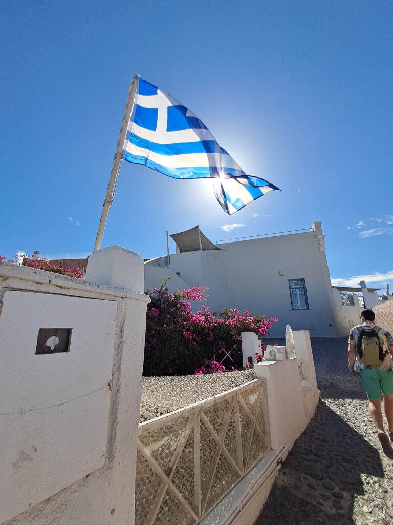 Walking through Oia, Santorini, past a Greek flag on a cobbled street, before heading off to explore by car