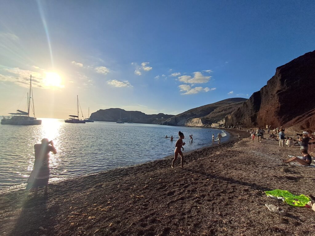 Golden hour at a quiet cove in Santorini, boats anchored offshore and people swimming, one of the spots only reachable easily by car