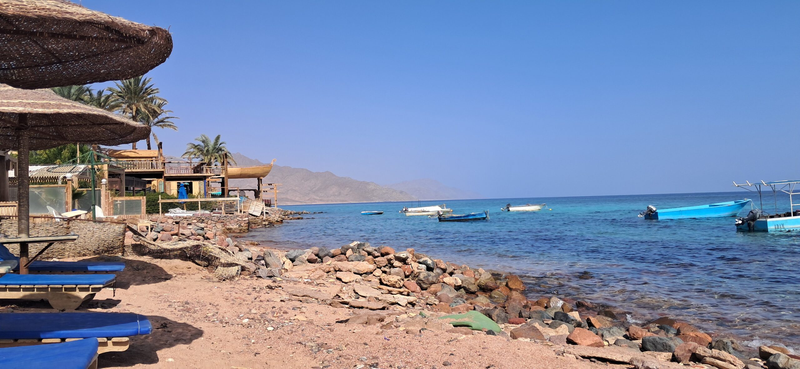 The rocky Dahab seafront meeting the turquoise Red Sea, with boats and desert mountains behind, on a calm January day