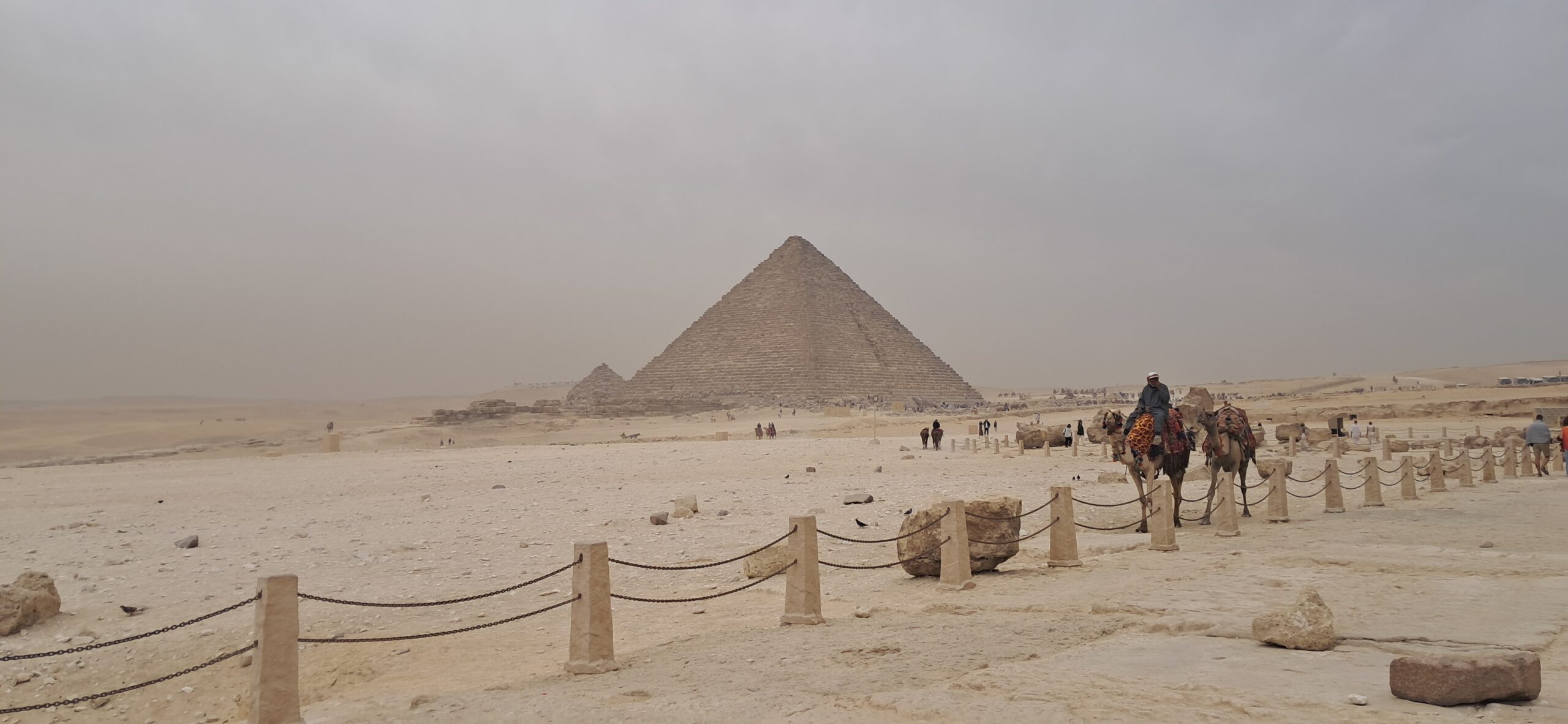 A camel and its handler by a rope barrier at the Pyramids of Giza on a hazy January morning, a pyramid rising behind through the haze