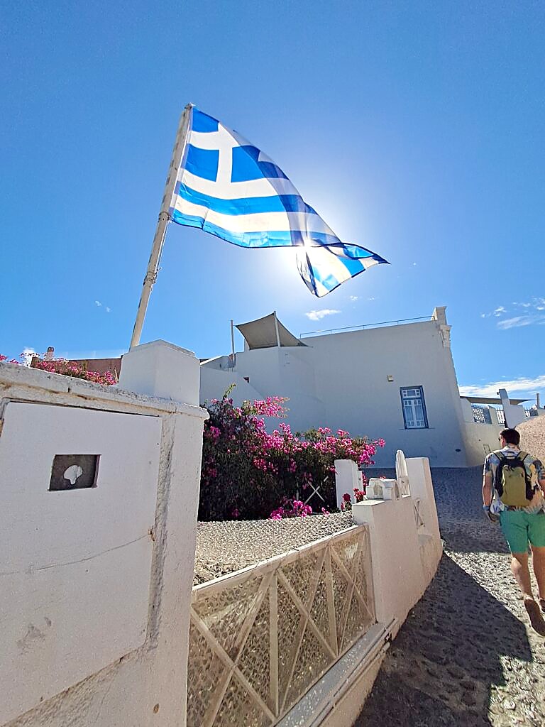 Walking through Oia, Santorini, past a Greek flag on a cobbled street, before heading off to explore by car