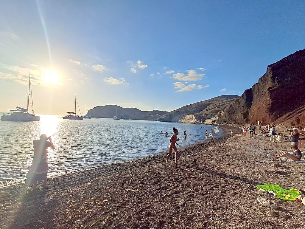 Golden hour at a quiet cove in Santorini, boats anchored offshore and people swimming, one of the spots only reachable easily by car